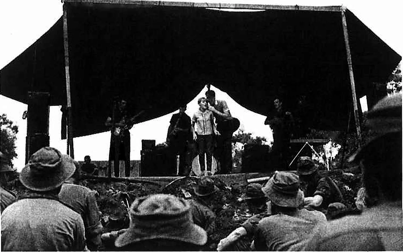 Troops seated on the ground watching Little Pattie and Col Joye perform under an open-sided stage